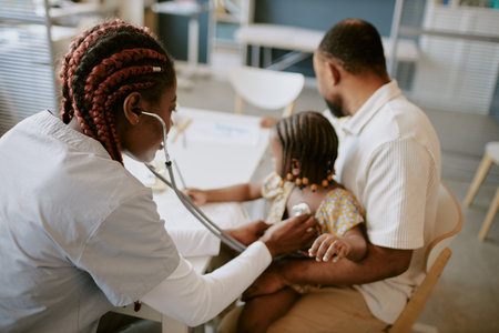 Female doctor using stethoscope for examining young girl while she sits on fathers lap in a clinic setting, focusing on health checkup and family careの写真素材