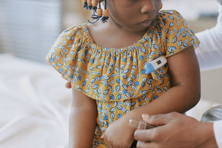 African American girl with arm thermometer sitting on bed while caregiver showing concern and providing comfort in softly lit room during healthcare check-upの写真素材