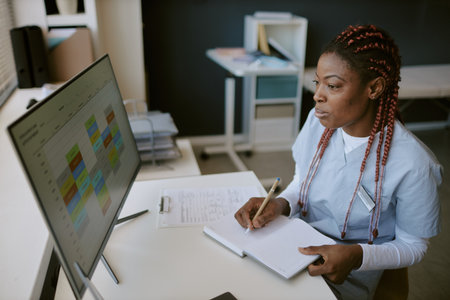 African American nurse in blue scrubs reviewing patient documents at desk in hospital environmentの写真素材