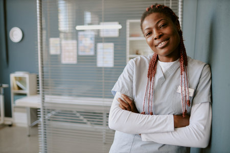 Portrait of smiling African American nurse leaning on wall in hospital corridor with arms crossed, wearing scrub uniform and standing confidentlyの写真素材