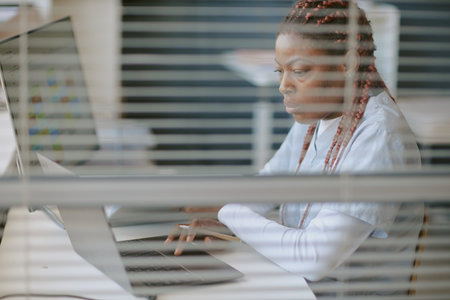 Black woman sitting at office desk, working on laptop with a focused expression, multiple screens visible in background, representing modern workspace without distractionsの写真素材
