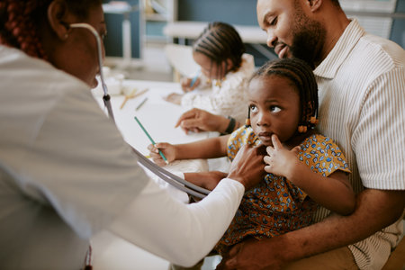Child sitting on fathers lap holding his hand while doctor examining her. African American family in...
