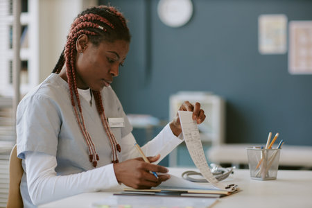 African American woman examining documents in medical setting, wearing scrubs, sitting at desk with medical tools, demonstrating focus and dedicationの写真素材