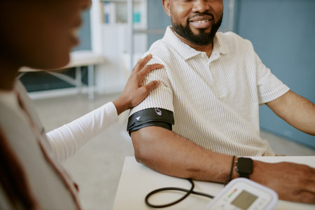 Smiling man sitting at medical examination table receiving blood pressure reading from a nurse in a clinical setting, with monitoring device and patient interaction presentの写真素材