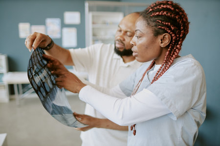 Two African American medical professionals examining X-rays in clinical setting, discussing patient health while wearing lab coats, with healthcare posters visible in backgroundの写真素材