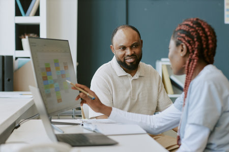 Two professionals discussing work tasks at an office desk while viewing digital charts on a monitor Clients or colleagues collaborating and sharing ideas in workplaceの写真素材