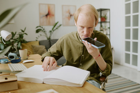 Mature Caucasian man suffering from visual impairment recording voice message while sitting at table...