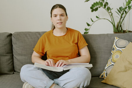 Young Brunette woman resting on comfortable sofa and enjoying novel written in Braille writing...