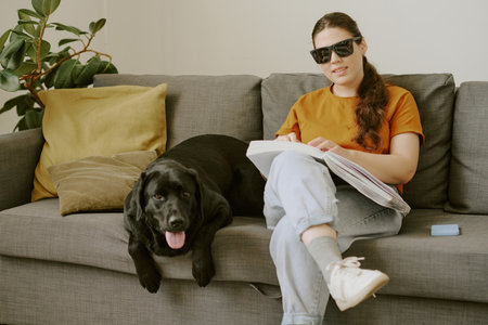Young woman with disability sitting on sofa and reading Braille book while her pet lying next to herの写真素材