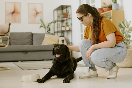 Young woman petting excited black Labrador while he lying on floorの写真素材