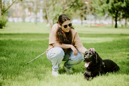 Young brunette woman with disability petting her lovely black Labrador while he lying on grass and restingの写真素材