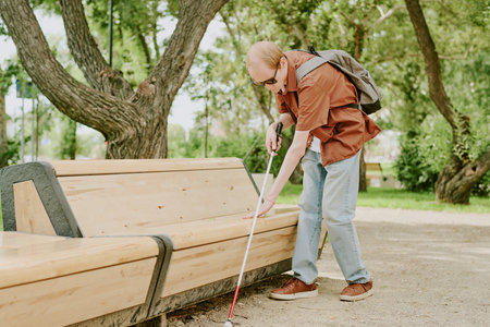 Caucasian blind man using metal cane to walk in park alone, he touching benchの写真素材