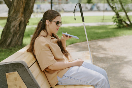 Side view shot of young woman recording voice message in social network while resting on bench in parkの写真素材