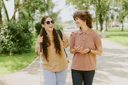 Two best friends laughing out loud while walking in green park, brunette young woman using cane to orientateの写真素材