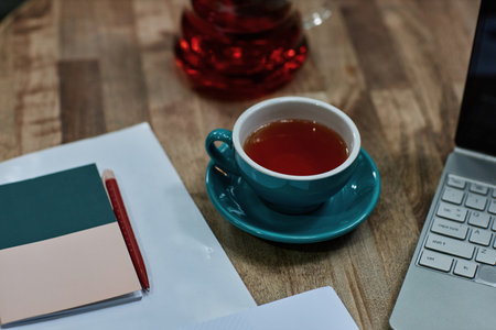 High angle shot of turquoise ceramic cup of black tea on wooden table with laptop and notes