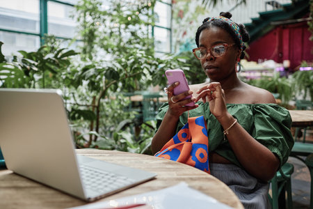 Famous African American female blogger checking social network while spending time in eco cafe with green plantsの写真素材