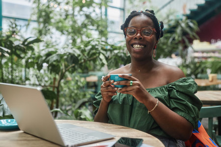 Smiling African American woman holding ceramic cup of tea, she looking at camera while sitting at wooden tableの写真素材