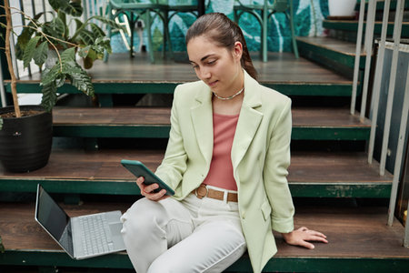 High angle shot of young Caucasian woman chatting on social network while having break in eco cafe, she resting on stairsの写真素材