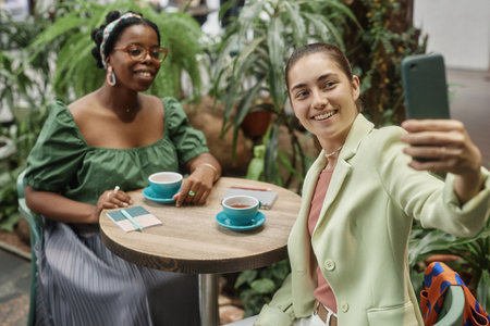 High angle shot of Caucasian young woman taking selfie with her African American friend while they enjoying black tea in eco cafeの写真素材