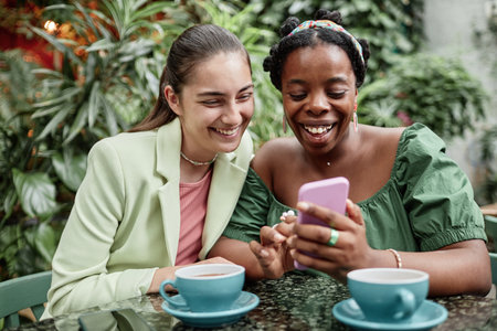Biracial besties enjoying social media while resting in eco cafe togetherの写真素材