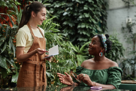 Smiling Caucasian waitress taking order in eco cafe, smiling female African American customer sitting at table and telling her orderの写真素材