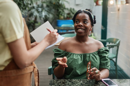 African American woman sitting at table and smiling while making order in cafe, unrecognizable waitress standing next to table and taking notesの写真素材