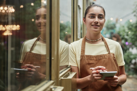 Young Caucasian waitress smiling lightly and leaning on golden door while posing for portraitの写真素材