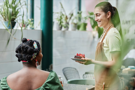 Smiling Caucasian waitress holding plate with chocolate cake while unrecognizable customer waiting for dessertの写真素材