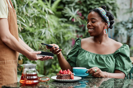 Beaming African American customer tapping smartphone to pay for food and beverage in eco cafeの写真素材