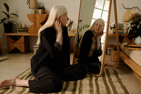 Young gen Z girl with albinism sitting on floor in living room and looking in mirror embracing her unique beauty and expressing self-loveの写真素材