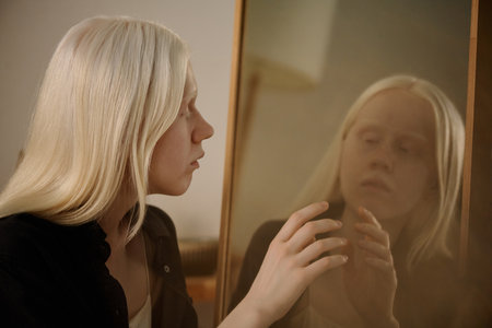 Medium closeup of mysterious young Caucasian woman with albinism looking in mirror and touching her reflectionの写真素材