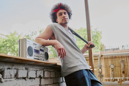 Low angle shot of young curly haired man holding wooden bat and leaning on old gray bricksの写真素材