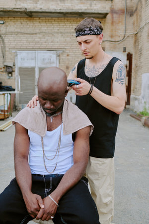 Young Caucasian man with tattoos shaving friends occiput with help of electric trimmer in old backyardの写真素材