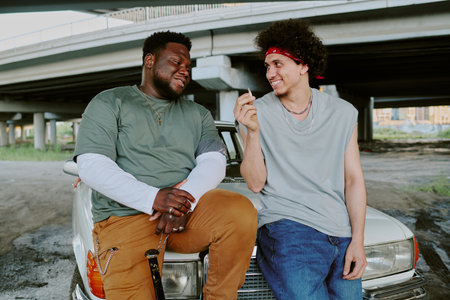 Biracial best friends smiling to each other while curly haired one smoking cigarette, they leaning on retro carの写真素材
