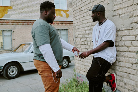 African American man giving wad of cash to guy who leaning on old garageの写真素材