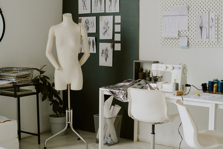 Creamy colored mannequin standing next to white table with metallic piece of fabric and white sewing machineの写真素材