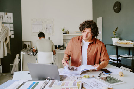 Fashionable designer turning over album pages with clothing sketches while tailor sitting at table and using sewing machine in backgroundの写真素材