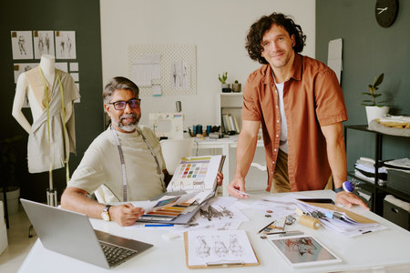Portrait of mature designer with album in hands sitting at work desk and tailor standing next to desk while posing for photoの写真素材