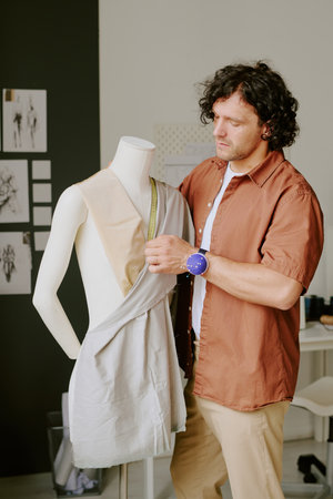 Caucasian curly haired tailor standing next to white plastic mannequin and measuring cotton fabricの写真素材