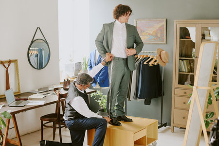 Tailor standing on one knee and doing measurements while curly haired model looking in mirrorの写真素材