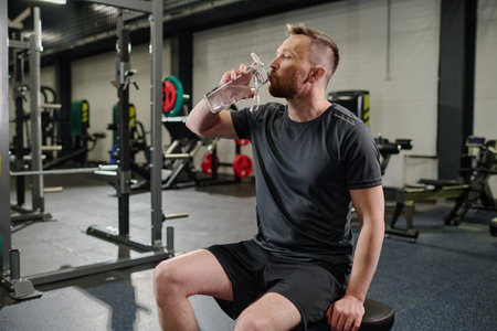 Shredded man sitting on sport bench and drinking pure waterの写真素材