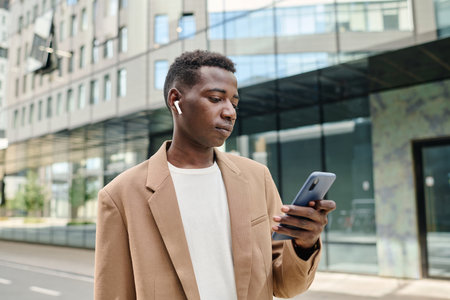 Gen Z Black man standing outdoors in modern business district changing playlist on smartphone on way to workの写真素材