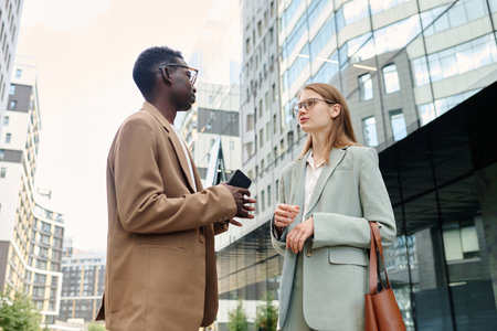 Low angle view shot of young Black and Caucasian coworkers wearing stylish formal clothes standing outdoors and having conversationの写真素材