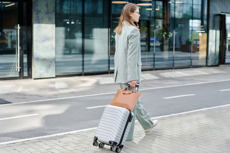Wide shot of young Caucasian woman walking along business district street with her luggage and talking on phoneの写真素材