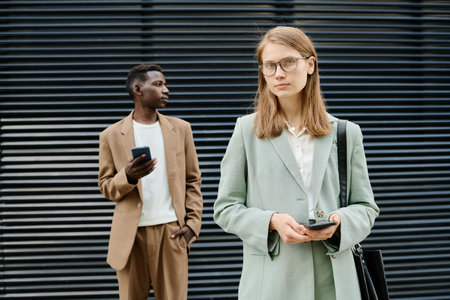 Medium portrait of young Caucasian female manager holding smartphone and looking at camera, her Black male colleague standing relaxed in backgroundの写真素材