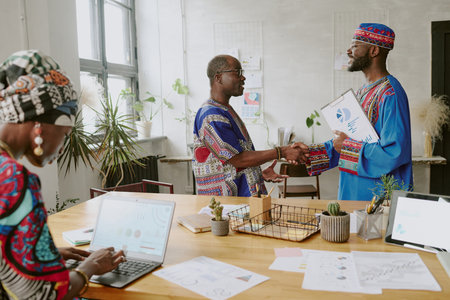Mature African American boss thanking his subordinate for successful project and giving handshake while female subordinate making presentation in foregroundの写真素材