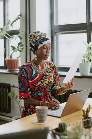 Vertical shot of African American woman sitting at work desk and reading report in modern officeの写真素材