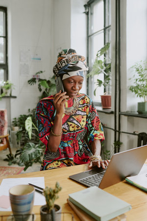 Vertical shot of African American worker using laptop while speaking on phone in officeの写真素材