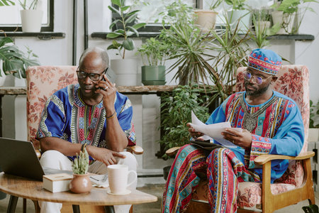 Mature African American worker speaking on phone while his colleague reading report during break timeの写真素材