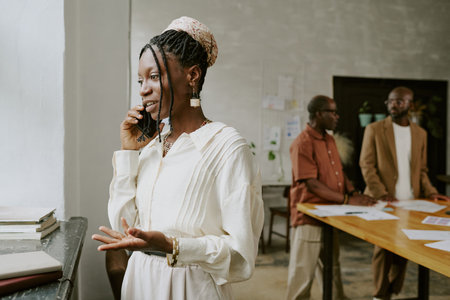 African American female office worker speaking on phone with client while her male colleagues discussing analytical report in backgroundの写真素材